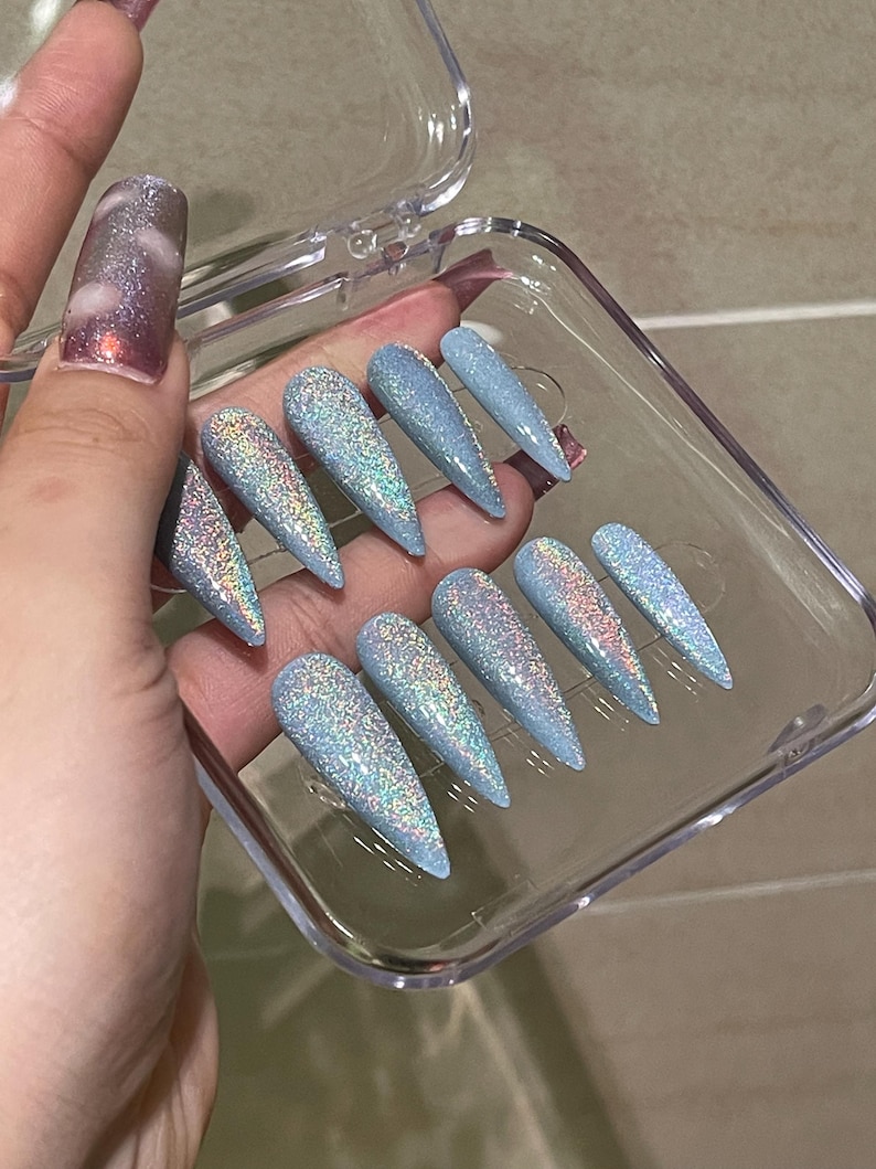 K&ouml;nnte beinhalten: Ein Set aus zehn stilettof&ouml;rmigen Press-on-N&auml;geln in Hellblau mit einem dichten, mehrfarbigen holografischen Glitzer-Finish. Die N&auml;gel sind in einem durchsichtigen Kunststoffbeh&auml;lter angeordnet und weisen eine scharfe, spitz zulaufende Form auf.