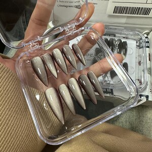 Peut inclure: Un &eacute;tui en plastique transparent contenant dix faux ongles longs et pointus. Les ongles sont de couleur brun iris&eacute; chatoyant. L'&eacute;tui est ouvert et les ongles sont visibles. Le texte "Taupe Brown 70015" est &eacute;crit sur une &eacute;tiquette.