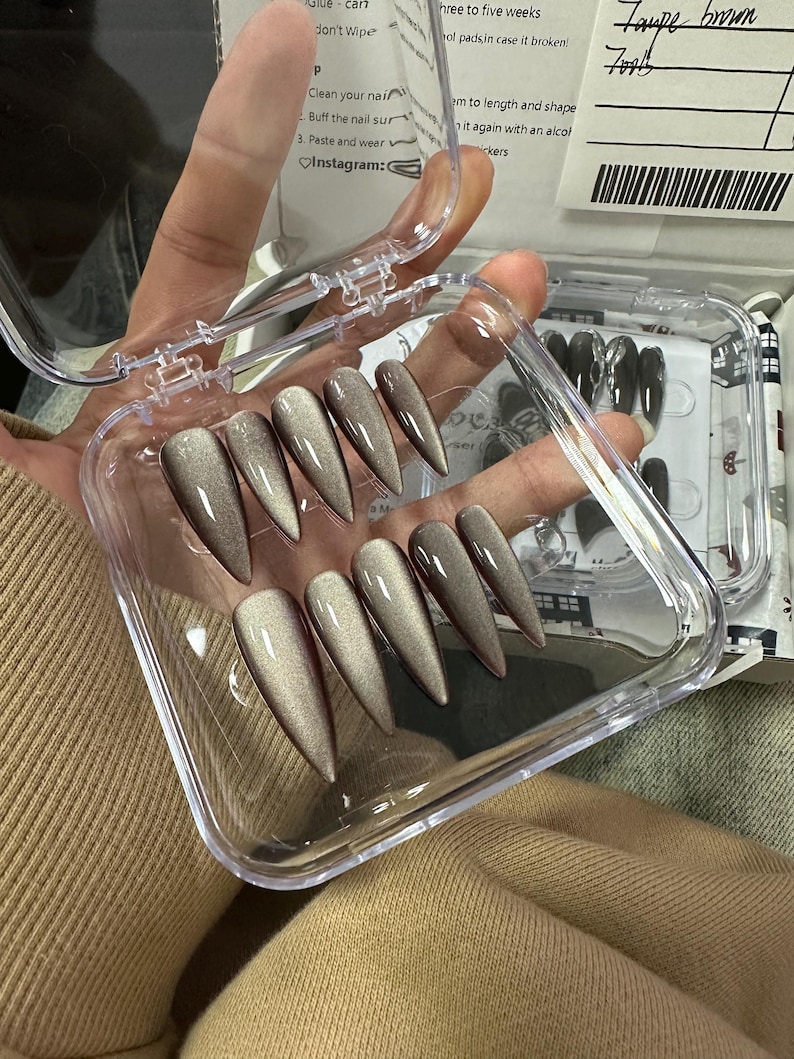 Peut inclure: Un &eacute;tui en plastique transparent contenant dix faux ongles longs et pointus. Les ongles sont de couleur brun iris&eacute; chatoyant. L'&eacute;tui est ouvert et les ongles sont visibles. Le texte "Taupe Brown 70015" est &eacute;crit sur une &eacute;tiquette.