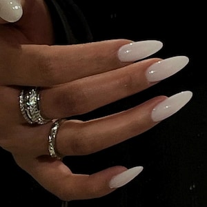 May include: Close-up of a hand with long, white almond-shaped nails. The nails are painted a milky white color and have a subtle shine. The hand is wearing two silver rings.