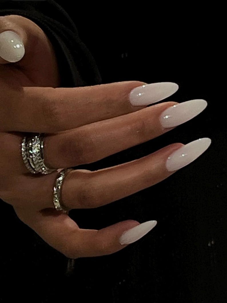 May include: Close-up of a hand with long, white almond-shaped nails. The nails are painted a milky white color and have a subtle shine. The hand is wearing two silver rings.