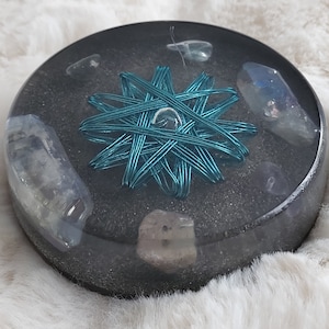 Puede incluir: Un posavasos redondo de resina transparente con una estrella de alambre turquesa en el centro. El posavasos está lleno de arena negra y varios cristales transparentes y opacos.