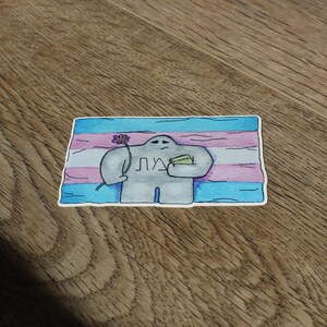 May include: A sticker featuring a cartoon figure holding a flower and a small green rectangle, set against a transgender pride flag background. The figure is gray, and the flag has blue, pink, and white stripes. The sticker has the word "ЛД" on it.