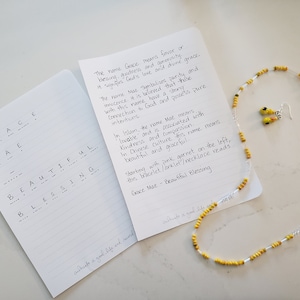 Puede incluir: Un conjunto de joyas hechas a mano con un collar y pendientes con cuentas amarillas y plateadas. El conjunto se muestra con dos cuadernos abiertos con texto manuscrito sobre el nombre "Grace Mae" y su significado.