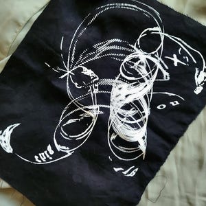 Può includere: Toppa in tessuto nero con un disegno astratto bianco e la scritta "birds" stampata sopra. Una piccola scultura bianca con corna è posta sopra la toppa. La toppa è su una superficie verde chiaro.