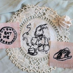Könnte beinhalten: Eine skurrile Anordnung mit einer cremefarbenen Deckchen, drei Stoffquadraten mit schwarzen Illustrationen und einem kleinen, lächelnden, weißen Keramikgesicht. Auf einem Quadrat steht "EAT ME". Das zentrale Bild zeigt eine kaninchenartige Figur mit einer Teetasse.