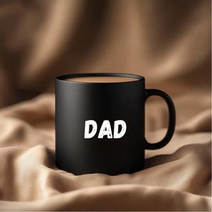 May include: Black ceramic coffee mug with the word "DAD" printed in white on the front. The mug is filled with coffee and is sitting on a brown surface.