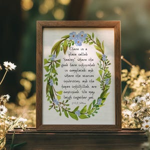 May include: A framed watercolour print with a wooden frame, featuring a quote surrounded by a floral wreath. The text reads "There is a place called 'heaven' where the good here unfinished is completed..." The print is displayed on a wooden crate.