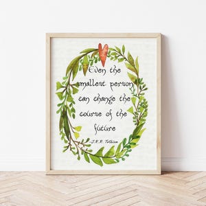 Op de afbeelding: Een ingelijste aquarelprint met een citaat van J.R.R. Tolkien: "Zelfs de kleinste persoon kan de loop van de toekomst veranderen." Het citaat is omgeven door een groene en oranje bloemenkrans. De lijst heeft een lichte houtkleur.