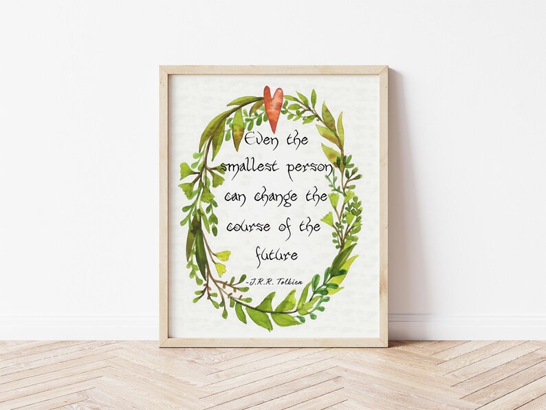 Op de afbeelding: Een ingelijste aquarelprint met een citaat van J.R.R. Tolkien: "Zelfs de kleinste persoon kan de loop van de toekomst veranderen." Het citaat is omgeven door een groene en oranje bloemenkrans. De lijst heeft een lichte houtkleur.