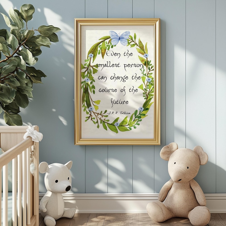 Op de afbeelding: Een ingelijste print met een gouden rand met de quote "Zelfs de kleinste persoon kan de loop van de toekomst veranderen" van J.R.R. Tolkien. De tekst is omgeven door een groene krans met een blauwe vlinder en kleine blauwe bloemen.