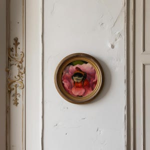 Può includere: Dipinto rotondo incorniciato di un'ape su un fiore rosa. Il dipinto ha una cornice color oro ed è montato su una parete bianca. L'ape è nera e gialla, e il fiore è di un rosa vibrante.
