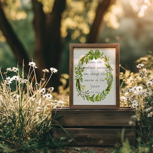 Op de afbeelding: Een ingelijste print met de quote: "Zelfs de kleinste persoon kan de loop van de toekomst veranderen." Het kunstwerk toont een groene krans met klavers en bladeren. De lijst is bruin en de print staat op een houten kist te midden van bloemen.