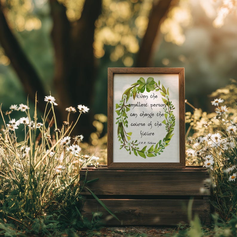 Op de afbeelding: Een ingelijste print met de quote: "Zelfs de kleinste persoon kan de loop van de toekomst veranderen." Het kunstwerk toont een groene krans met klavers en bladeren. De lijst is bruin en de print staat op een houten kist te midden van bloemen.