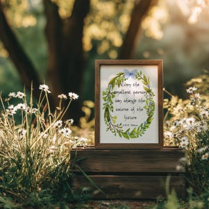 Op de afbeelding: Een ingelijste print met de quote: "Zelfs de kleinste persoon kan de loop van de toekomst veranderen." Het kunstwerk toont een groene krans met blauwe bloemen en een vlinder. De lijst is bruin en de print staat op een houten krat.