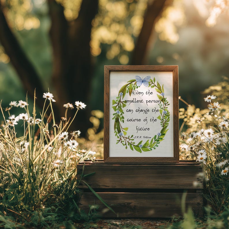 Op de afbeelding: Een ingelijste print met de quote: "Zelfs de kleinste persoon kan de loop van de toekomst veranderen." Het kunstwerk toont een groene krans met blauwe bloemen en een vlinder. De lijst is bruin en de print staat op een houten krat.