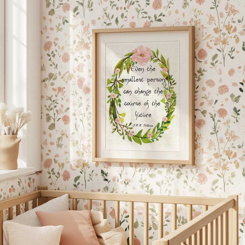 Op de afbeelding: Een ingelijste print met een bloemenkrans en de quote "Zelfs de kleinste persoon kan de loop van de toekomst veranderen" van J.R.R. Tolkien. De print zit in een licht houten frame, tegen een bloemenbehang.