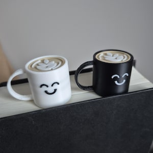 May include: Two small ceramic coffee mugs with smiling faces. One mug is white with a brown coffee design and the other is black with a white coffee design.