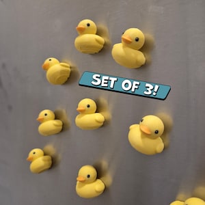 May include: A collection of yellow rubber duck magnets with orange beaks and black eyes. The text "SET OF 3!" is displayed on a blue banner. The magnets are attached to a metallic surface.