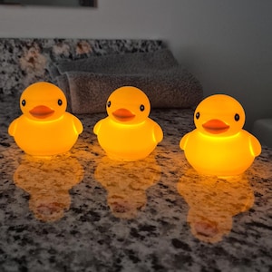 May include: Three glowing yellow rubber ducks with orange beaks and black eyes. Each duck emits a warm, internal light, creating a soft illumination that reflects on the surface below. The ducks are positioned in a row, displaying a smooth, rounded form.