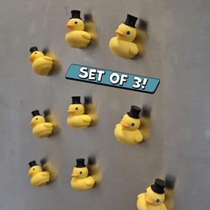 Peut inclure: Un ensemble de trois aimants canard en caoutchouc jaune, chacun portant un chapeau haut de forme noir. Les canards ont des becs orange et sont disposés sur une surface métallique. Le texte "SET OF 3!" est affiché dans une bannière bleue.