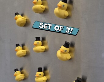 Puede incluir: Un conjunto de tres imanes de pato de goma amarillos, cada uno con un sombrero de copa negro. Los patos tienen picos naranjas y están colocados sobre una superficie metálica. El texto "SET OF 3!" se muestra en una pancarta azul.