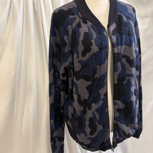 K&ouml;nnte beinhalten: Eine lang&auml;rmelige Bomberjacke mit Camouflage-Muster in Marineblau, Anthrazit und Schwarz. Die Jacke verf&uuml;gt &uuml;ber einen einfarbigen schwarzen Rippstrickkragen, B&uuml;ndchen und Saum sowie einen durchgehenden silberfarbenen Metallrei&szlig;verschluss vorne.