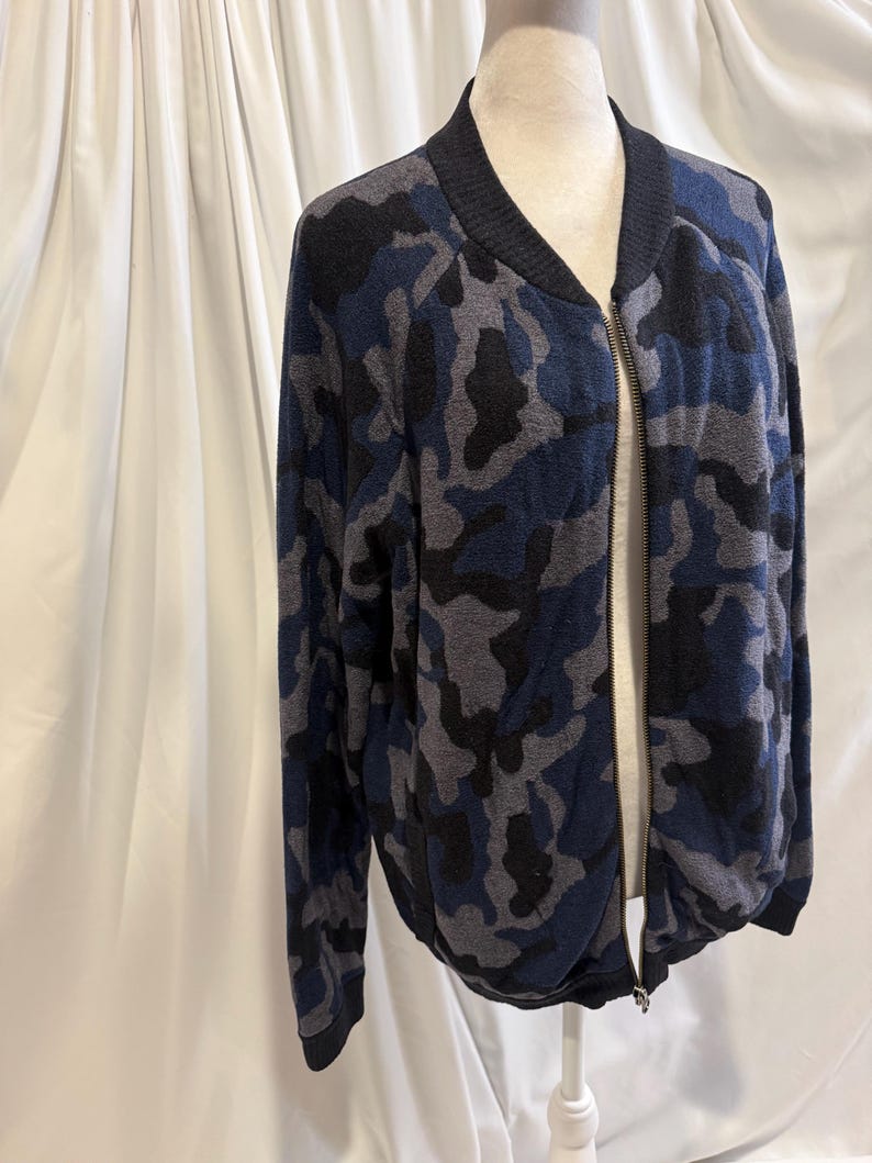 K&ouml;nnte beinhalten: Eine lang&auml;rmelige Bomberjacke mit Camouflage-Muster in Marineblau, Anthrazit und Schwarz. Die Jacke verf&uuml;gt &uuml;ber einen einfarbigen schwarzen Rippstrickkragen, B&uuml;ndchen und Saum sowie einen durchgehenden silberfarbenen Metallrei&szlig;verschluss vorne.