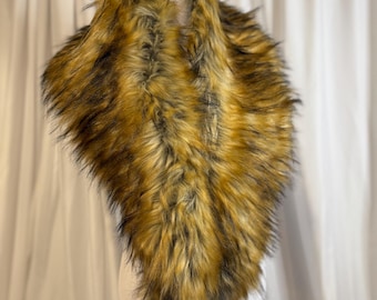 May include: A faux fur scarf in shades of brown, tan, and black, displayed on a white mannequin. The scarf has a fluffy, textured appearance, and is designed to be worn around the neck. The background is a white curtain.
