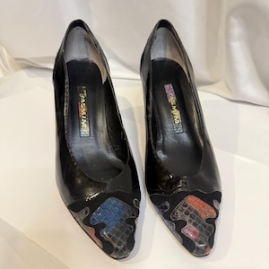 May include: A pair of black patent leather pumps with a pointed toe and a high heel. The toe features a snakeskin pattern in shades of blue, red, and gray. The brand name "JASMINE" is visible inside the shoe.
