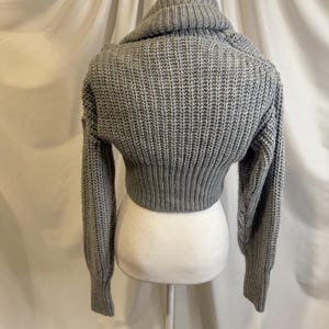 Puede incluir: Un su&eacute;ter corto gris con cuello alto. El su&eacute;ter est&aacute; hecho de una tela de punto acanalado grueso. Las mangas son largas y ajustadas, y el dobladillo inferior llega a la cintura. El su&eacute;ter se exhibe en un maniqu&iacute; blanco.