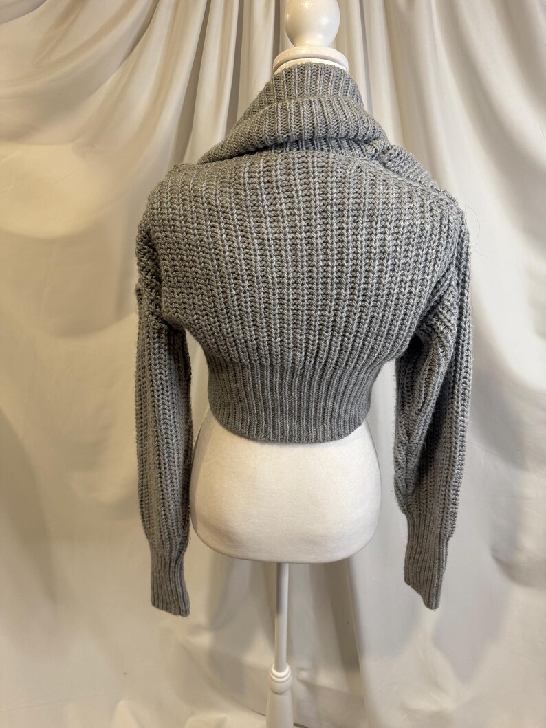 Puede incluir: Un su&eacute;ter corto gris con cuello alto. El su&eacute;ter est&aacute; hecho de una tela de punto acanalado grueso. Las mangas son largas y ajustadas, y el dobladillo inferior llega a la cintura. El su&eacute;ter se exhibe en un maniqu&iacute; blanco.