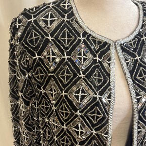 May include: A black jacket featuring an intricate geometric pattern created with silver sequins and white seed beads. The garment has a round neckline and long sleeves, with the silver and white beadwork forming a repeating diamond grid design.