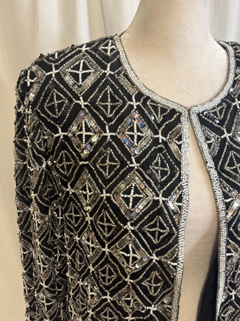 May include: A black jacket featuring an intricate geometric pattern created with silver sequins and white seed beads. The garment has a round neckline and long sleeves, with the silver and white beadwork forming a repeating diamond grid design.