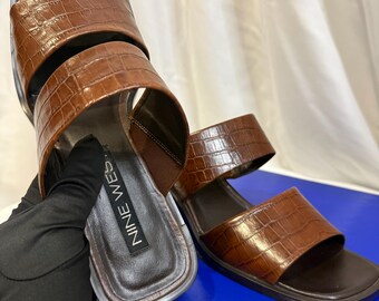 May include: Brown faux-leather sandals with a crocodile texture. The sandals have a square toe and two straps across the top of the foot. The brand name "NINE WEST" is visible on the insole. The sandals are displayed on a blue surface.