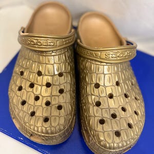 May include: A pair of metallic gold clogs featuring a textured crocodile skin pattern across the upper surface. Each shoe includes a pivoting heel strap with the brand name Crocs embossed on the side and multiple ventilation holes throughout.