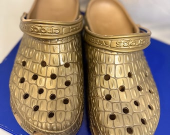 May include: A pair of metallic gold clogs featuring a textured crocodile skin pattern across the upper surface. Each shoe includes a pivoting heel strap with the brand name Crocs embossed on the side and multiple ventilation holes throughout.