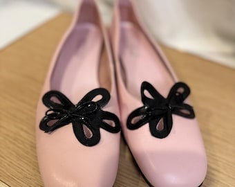 May include: A pair of light pink ballet flats with black flower-shaped embellishments. The shoes have a rounded toe and a low heel. The flats are displayed on a light-colored wooden surface.
