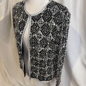 May include: A black long-sleeve jacket featuring an intricate geometric pattern of silver sequins and white beads. The open-front design includes a round neckline and is fully embellished with a repeating diamond grid motif across the entire surface.