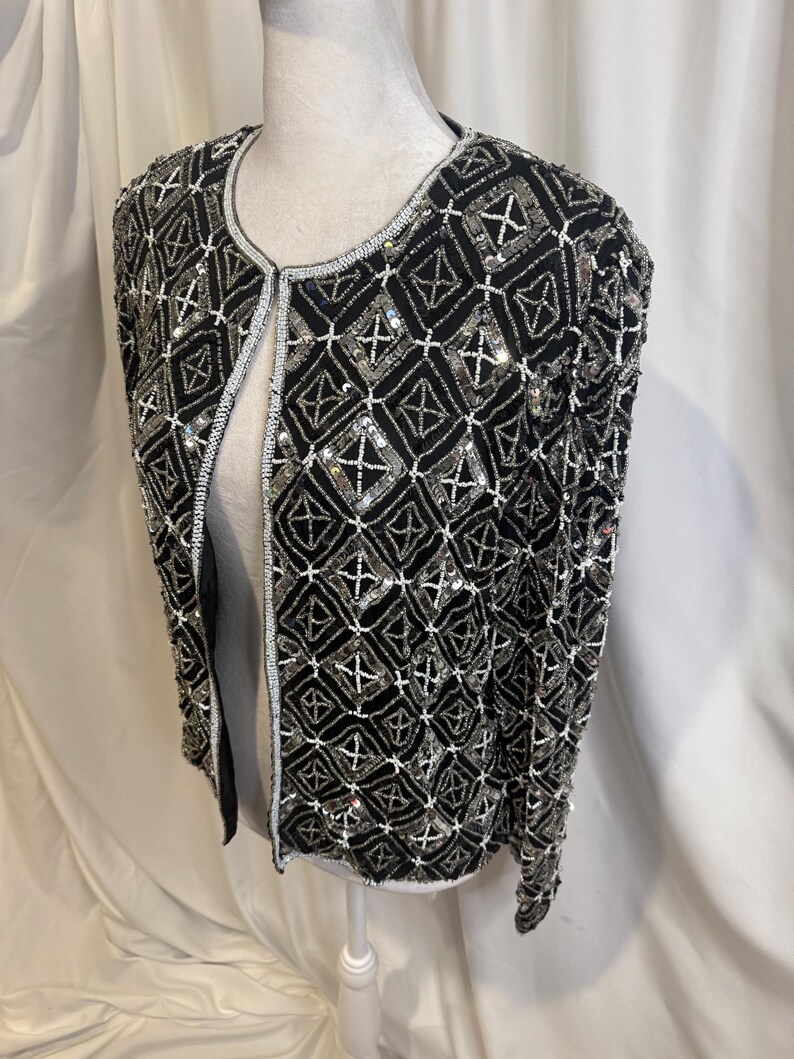 May include: A black long-sleeve jacket featuring an intricate geometric pattern of silver sequins and white beads. The open-front design includes a round neckline and is fully embellished with a repeating diamond grid motif across the entire surface.