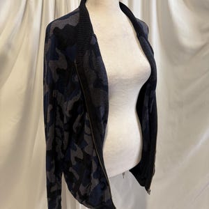K&ouml;nnte beinhalten: Eine lang&auml;rmelige Rei&szlig;verschlussjacke mit einem Tarnmuster in Dunkelblau, Schwarz und Grau. Das Kleidungsst&uuml;ck verf&uuml;gt &uuml;ber einen einfarbigen schwarzen Rippkragen und passende schwarze Rippb&uuml;ndchen sowie einen durchgehenden Frontrei&szlig;verschluss.
