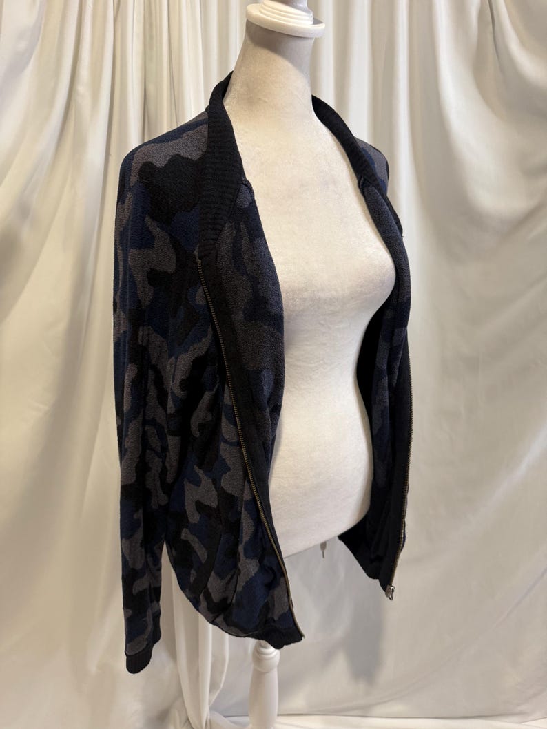 K&ouml;nnte beinhalten: Eine lang&auml;rmelige Rei&szlig;verschlussjacke mit einem Tarnmuster in Dunkelblau, Schwarz und Grau. Das Kleidungsst&uuml;ck verf&uuml;gt &uuml;ber einen einfarbigen schwarzen Rippkragen und passende schwarze Rippb&uuml;ndchen sowie einen durchgehenden Frontrei&szlig;verschluss.