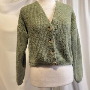 May include: A sage green cardigan with a V-neck and three large, round, light brown buttons. The textured knit fabric has a slightly fuzzy appearance. The sleeves are long, and the cardigan is displayed on a mannequin.