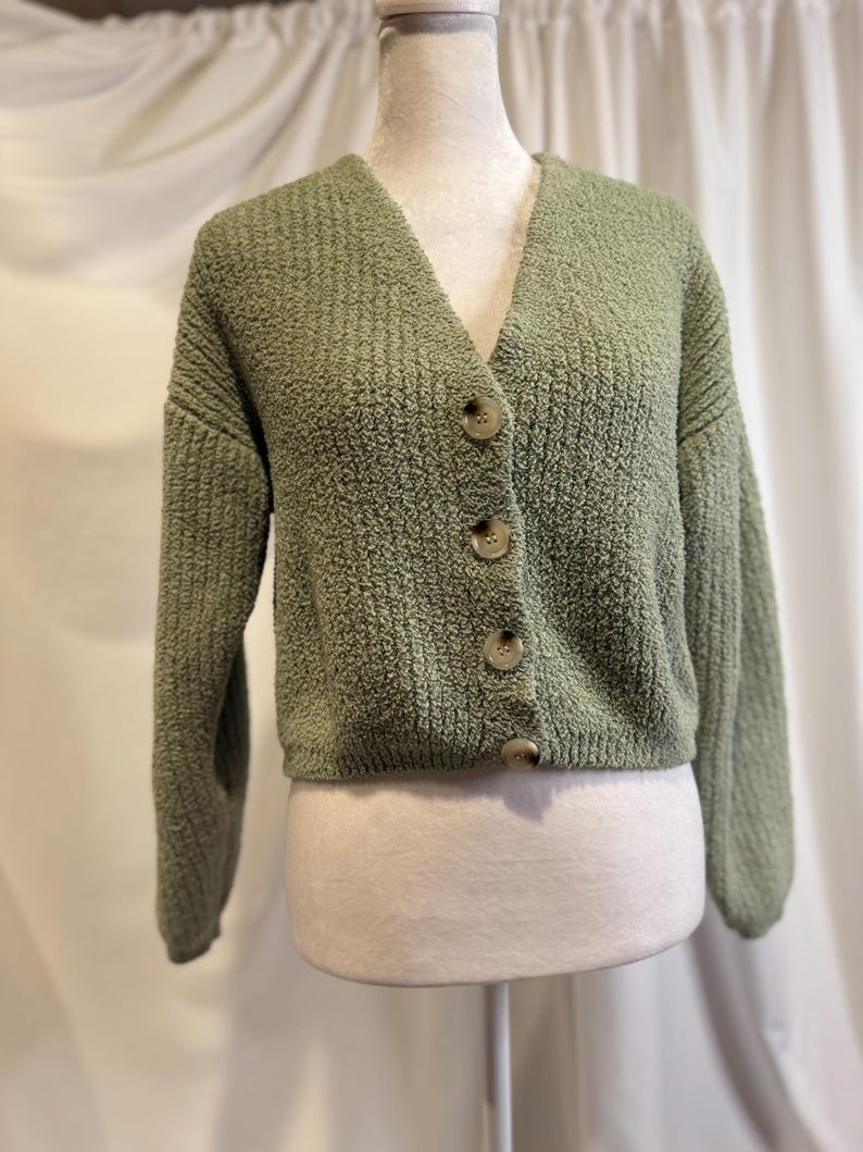 May include: A sage green cardigan with a V-neck and three large, round, light brown buttons. The textured knit fabric has a slightly fuzzy appearance. The sleeves are long, and the cardigan is displayed on a mannequin.