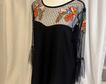 May include: Black dress with floral embroidery on a mannequin. The dress features a sheer, dotted overlay on the upper bodice and sleeves, with bell sleeves. The embroidery includes orange, red, and green floral designs.