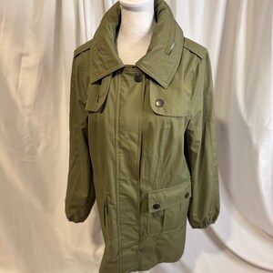 May include: Olive green jacket with a high collar, button closures, and two front pockets. The jacket is displayed on a white mannequin against a white backdrop. The jacket has a classic, utilitarian design.