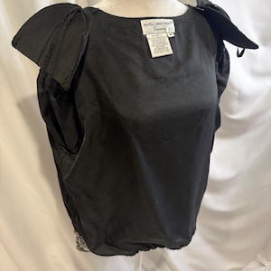 May include: A black blouse with short, structured puff sleeves and a round neckline. The garment features a label at the neck that reads Papell Boutique Evening, size M. The hem is gathered, and the fabric has a smooth, solid black finish.