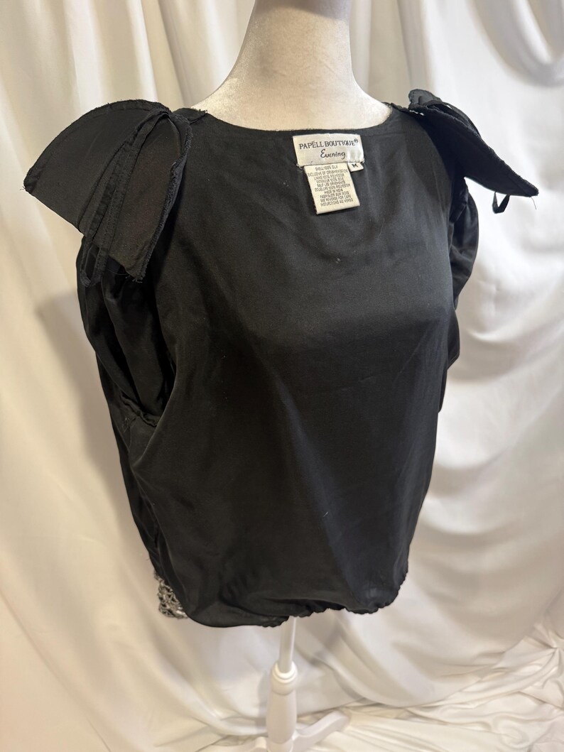 May include: A black blouse with short, structured puff sleeves and a round neckline. The garment features a label at the neck that reads Papell Boutique Evening, size M. The hem is gathered, and the fabric has a smooth, solid black finish.