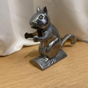 May include: A silver-colored metal squirrel nutcracker. The squirrel is standing upright on a log-shaped base, with its arms outstretched. The nutcracker has a detailed design, including the squirrel's fur and facial features.