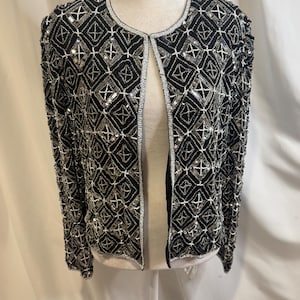 May include: A black open-front jacket featuring an intricate geometric pattern created with silver beads and sequins. The garment has long sleeves, a round neckline, and silver beaded trim along the edges, showcasing a dense, textured surface.